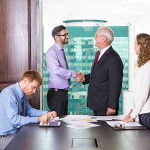 Smiling senior and young businessmen standing at head of table and shaking hands. Businesswoman is standing at table and looking at them. Another businessman is sitting at table and making notes.