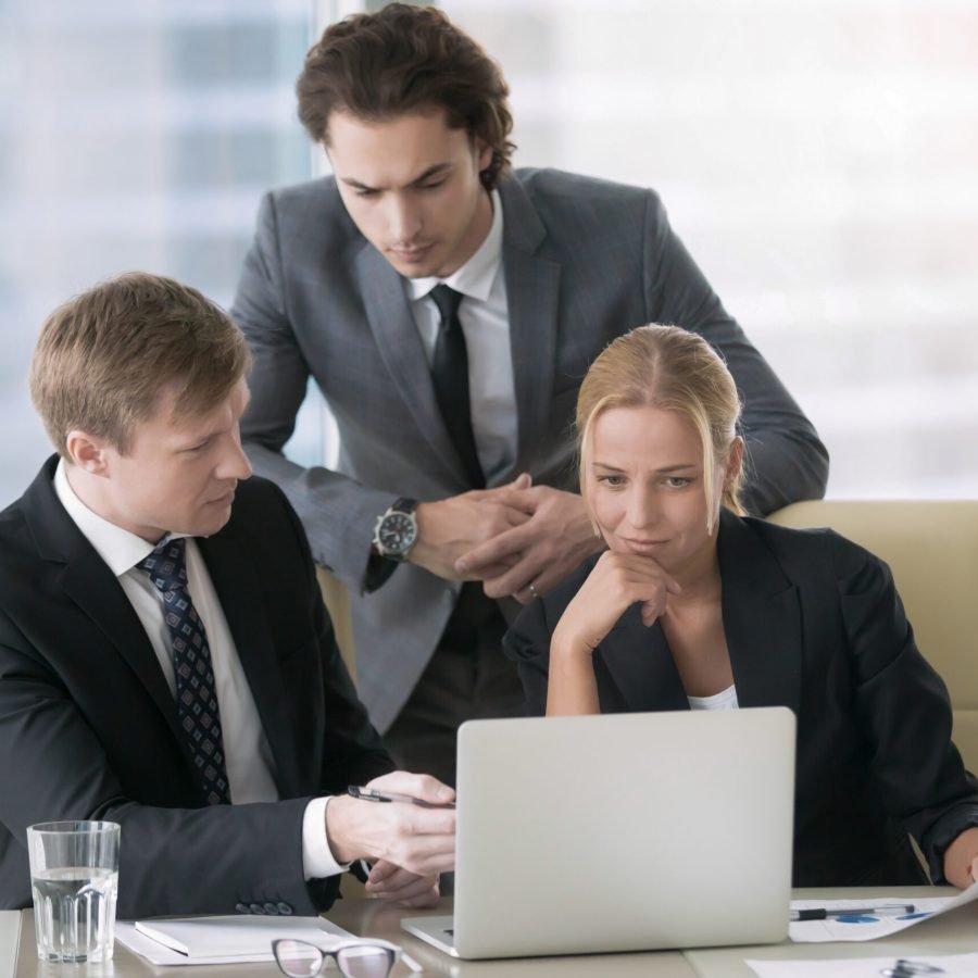 Group of three business partners discussing new project at meeting in office room, using laptop. Young confident businessman in formal wear suit explaining idea and showing presentation on screen